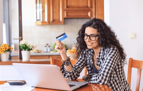 woman sitting in her kitchen in front of a laptop smiling while she holds up one of her credit cards