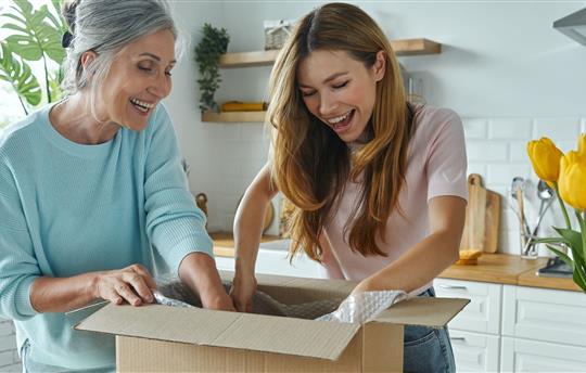 Happy senior woman and her adult daughter unpacking box while standing at the domestic kitchen