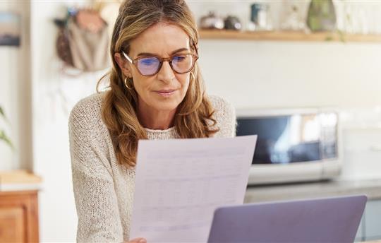 woman wearing glasses and looking at a printed document while sitting near her laptop