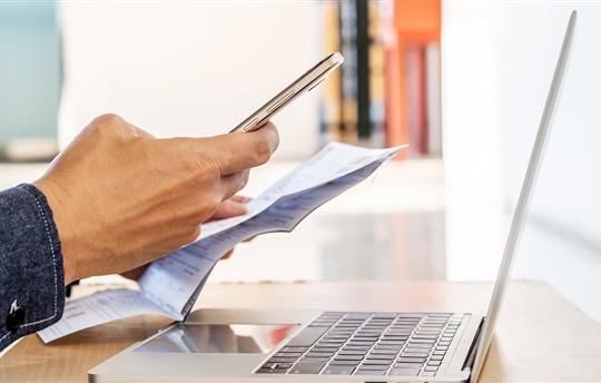 A person holding a mobile phone in one hand and a paper statement in the other while sitting in front of an open laptop