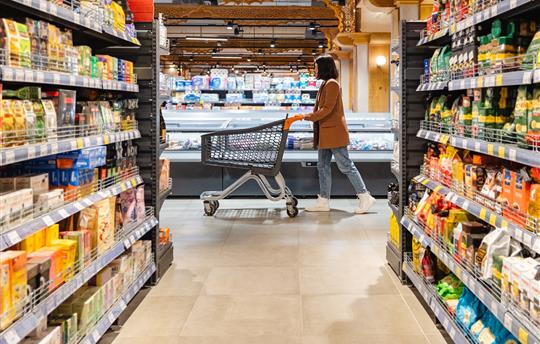 a woman with a cart walks between rows of shelves in a grocery store