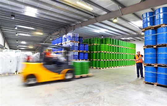 forklift and two workers in orange vests in a warehouse filled with blue and green drums stacked to the ceiling