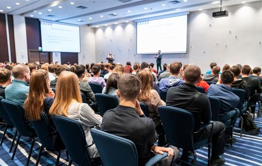 People sitting in a large conference room listening to a presenter in the front of the room