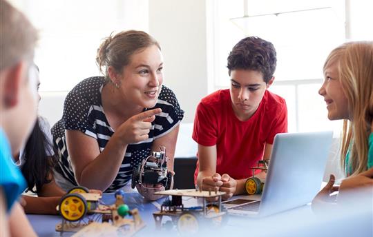 female teacher talking to four adolescent students while holding a small robot