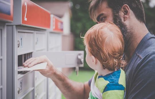 father holding infant son while collecting mail from a community mailbox