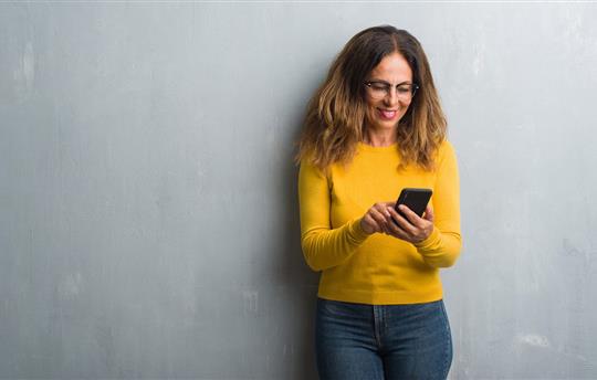 Middle aged woman in yellow sweater using smartphone standing and smiling