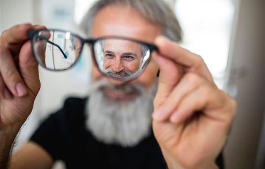 Older man with gray beard holding his eyeglasses out away from his face