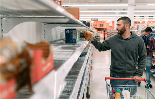 man shopping in large store, pushing a cart and looking at shelves with very little stock