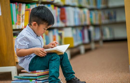 little boy sitting on a stack of books in the library and reading