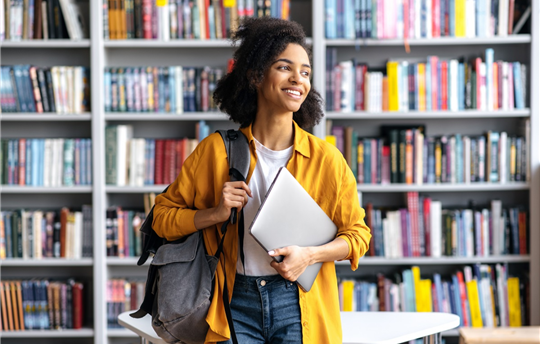 Portrait of a female student standing against background of bookshelves in university library holding laptop and backpack looking to the side