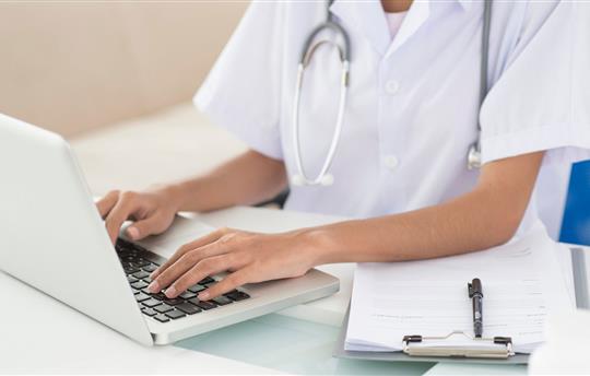 Doctor typing on laptop, clipboard with paperwork placed next to her