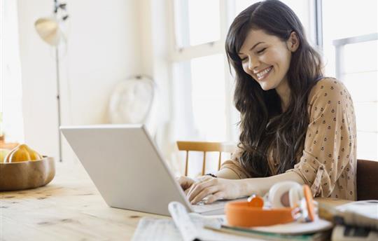 woman sitting at her dining room table, smiling while she views something on her laptop