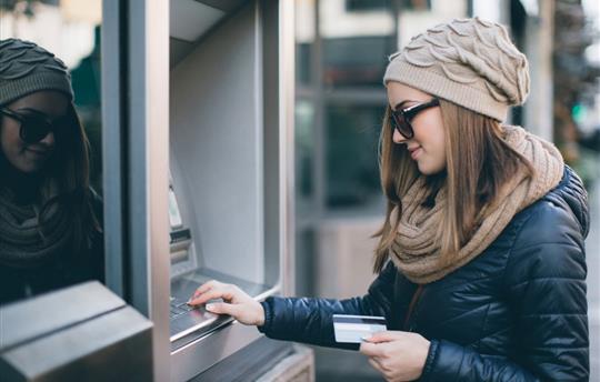 female millennial in winter coat and hat at an atm holding a debit card