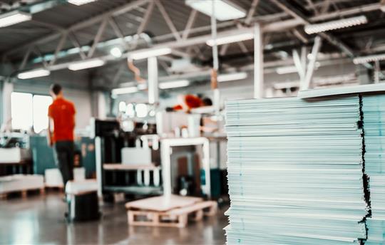 Piles of sheets in printing shop foreground, in background is a worker and printing machines