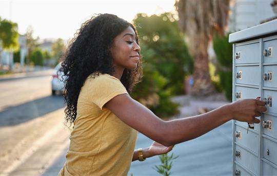 woman wearing yellow shirt and gold watch checking her mailbox near the curb