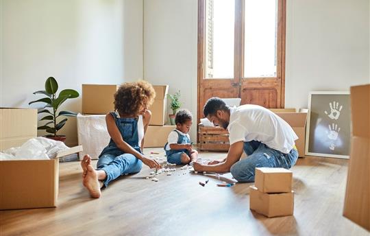 mom and dad with young child playing with small toys in the center of a room filled with moving boxes