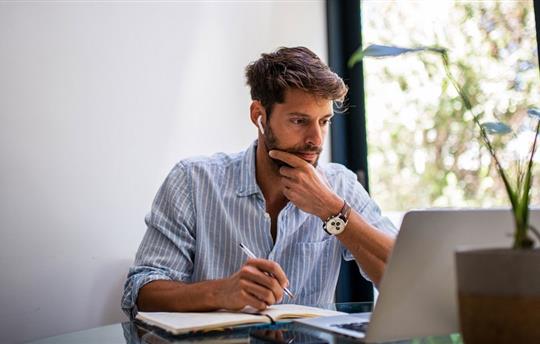 man working at home in front of his laptop, writing notes on a pad of paper