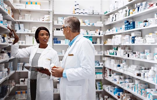 one female pharmacist and one male pharmacist in white coats talking to each other in pharmacy stock room