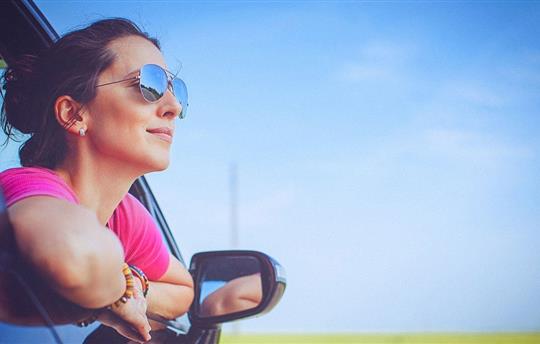 Woman in pink shirt and sunglasses, leaning out car window and smiling with blue sky in the background