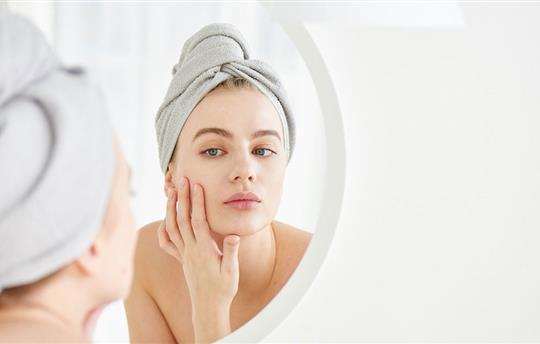 Portrait of young girl with towel on head in white bathroom looks and touches her face in the mirror