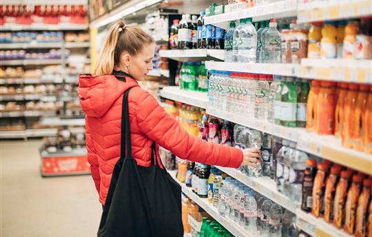 woman in puffy, orange jacket and black bag over her shoulder in an aisle at the grocery store looking at products
